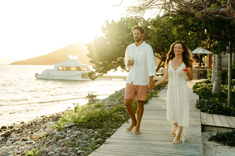 Couple walking on wooden boardwalk at sunset near beach with moored boat in background