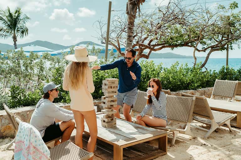 Four adults playing Jenga on a wooden table at a beachfront resort with ocean and islands visible