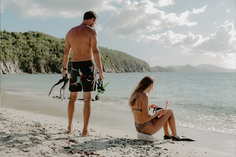 Two people with snorkeling gear standing in shallow ocean water near rocky cliffs on beach