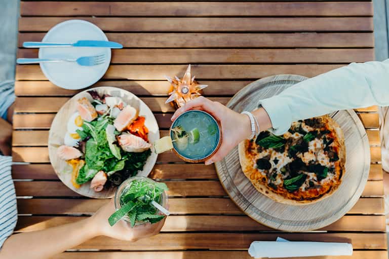Overhead view of waterfront dining table with seafood salad, pizza, and mojito cocktail on wooden deck