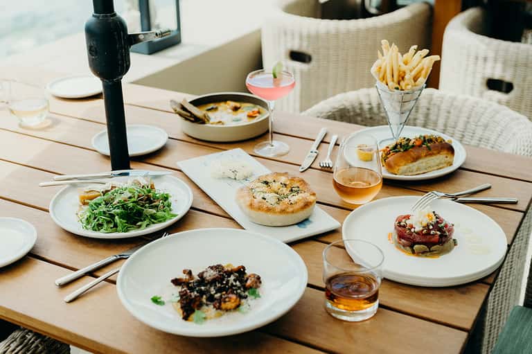 Outdoor dining table with gourmet dishes including salad, flatbread, seafood, fries, and cocktails in natural sunlight