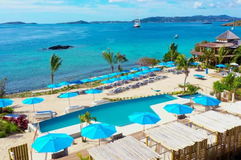 Luxury beachfront resort with turquoise pools, blue umbrellas, palm trees, and sailboats in the bay