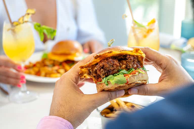 Hand holding a gourmet burger with fries and cocktails at an outdoor dining venue