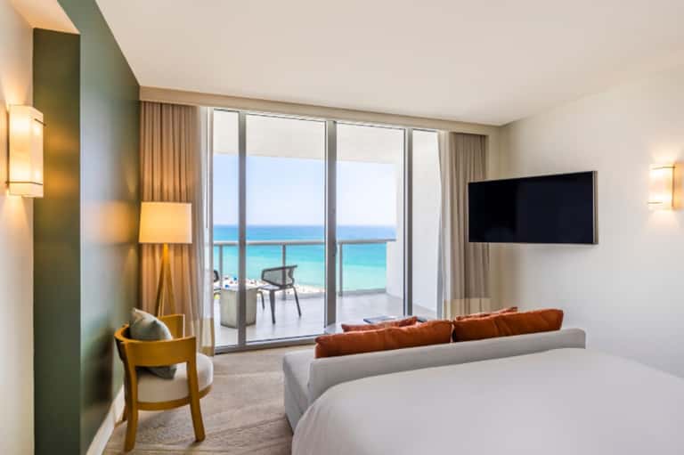 Luxury beachfront hotel room with ocean view, gray sofa, bed, and glass balcony doors opening to terrace