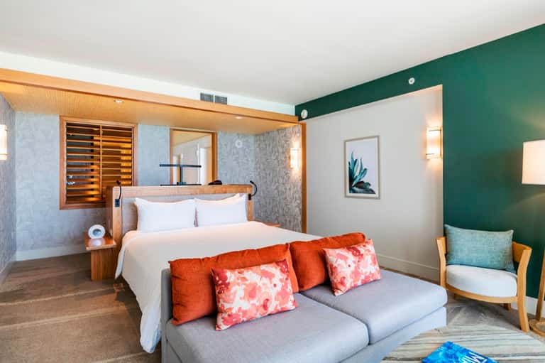 Modern hotel bedroom with green accent wall, wooden bed frame, and gray sofa with orange pillows