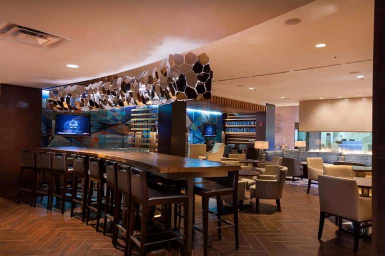 Hotel lounge with bar seating, geometric ceiling installation, wood flooring, and modern furnishings with blue accents