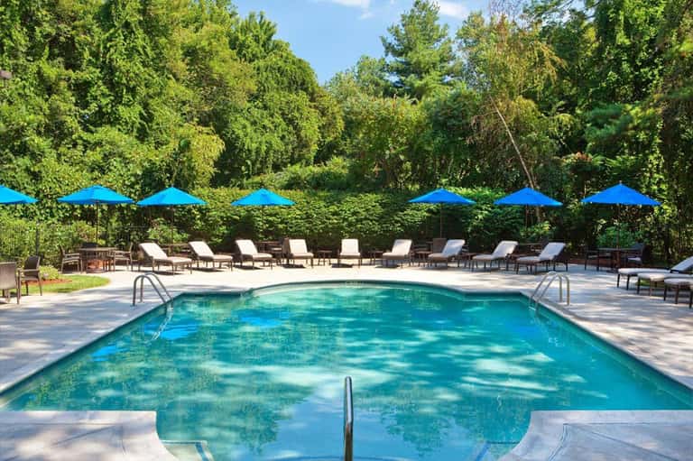 Resort pool with crystal blue water, lounge chairs, blue umbrellas, and lush green trees in background