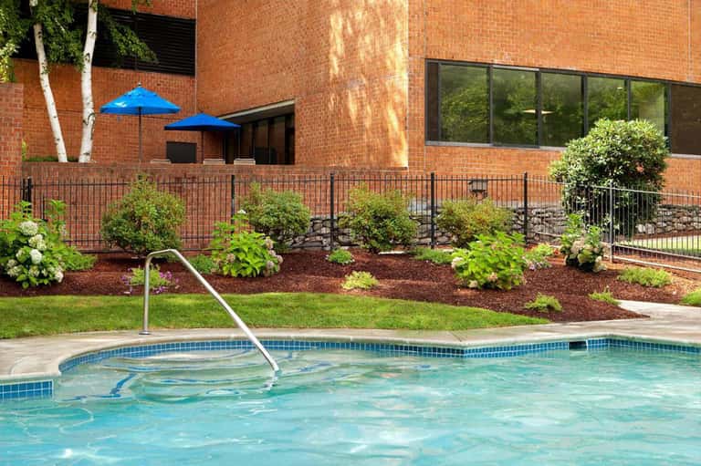 Resort pool with blue umbrellas, brick building, landscaped garden with shrubs and flowers