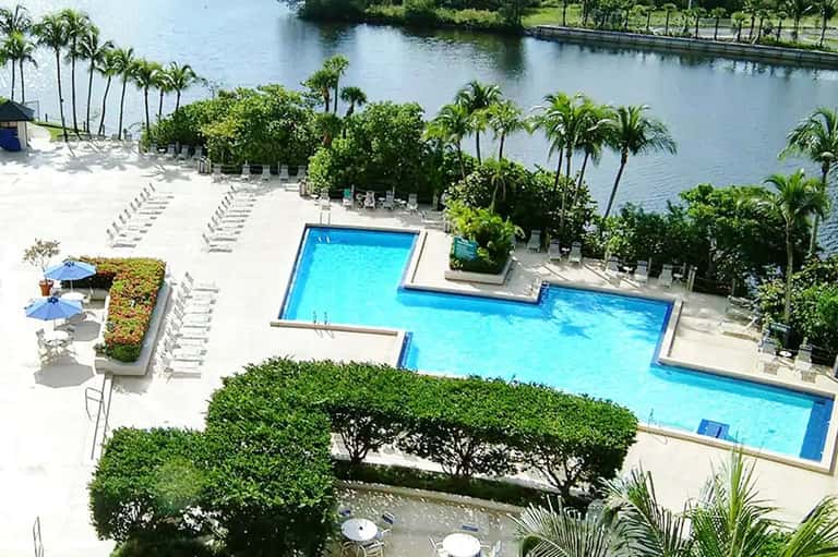 Aerial view of resort with multiple bright blue swimming pools, white loungers, palm trees, and waterfront along lagoon