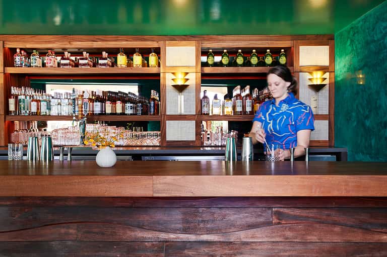 Bartender in blue patterned shirt preparing cocktails behind wooden bar with extensive liquor selection and green wall backdrop