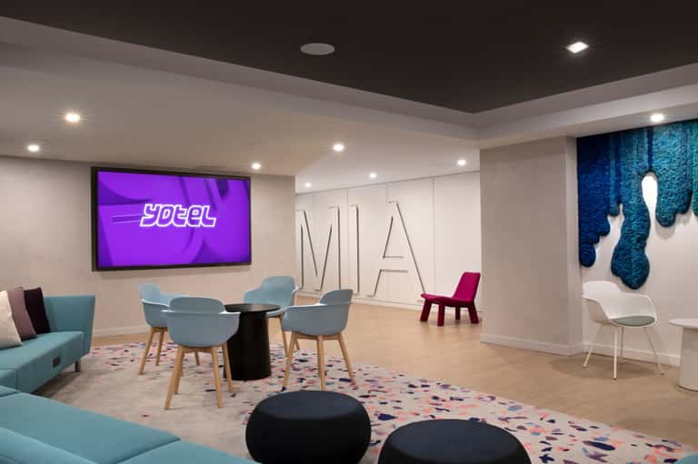 Modern hotel lobby with turquoise seating, black accent tables, and purple digital signage displaying 'YOTEL'