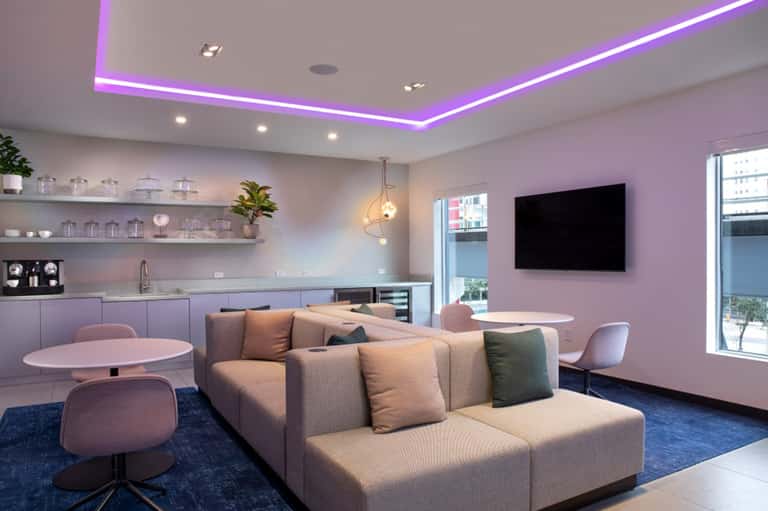 Modern lounge with pink walls, purple ceiling lighting, tan sofa, kitchenette, and round tables