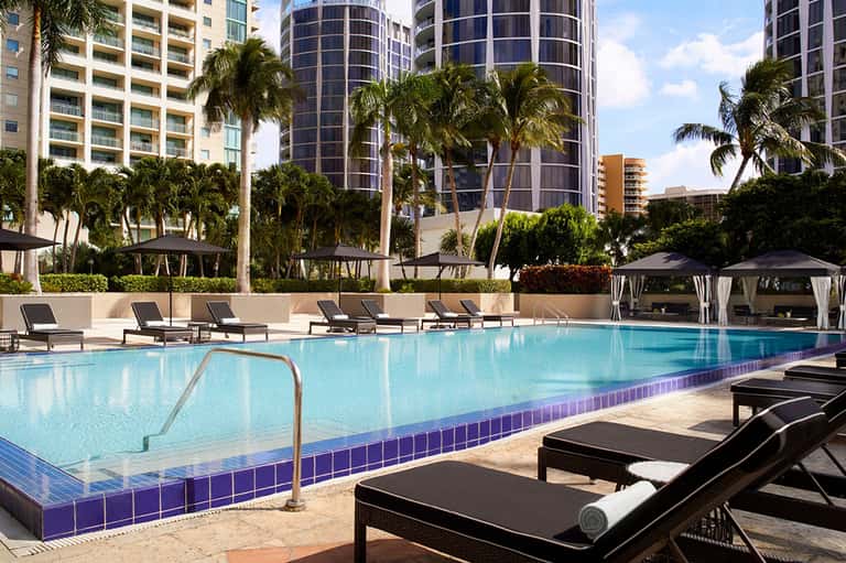 Luxury resort pool with turquoise water, black loungers, palm trees, and high-rise hotels in background
