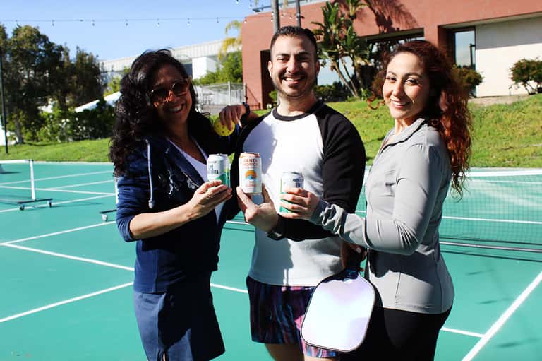 Three people holding beverages on a turquoise tennis court on a sunny day