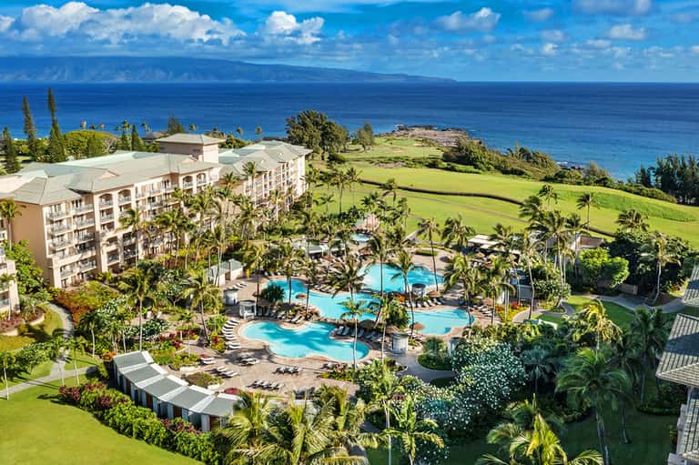 Aerial view of oceanfront resort with curved pools, palm trees, and manicured lawns overlooking blue ocean and distant island.