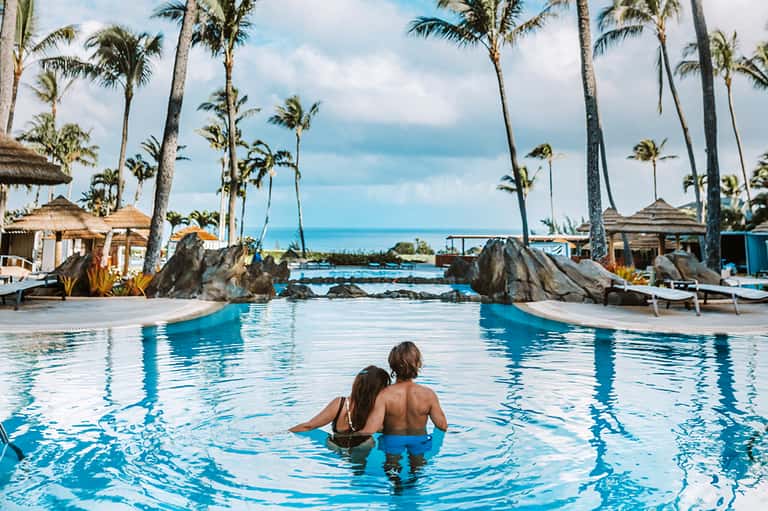 Two children wading in tropical resort pool with palm trees and ocean view in background