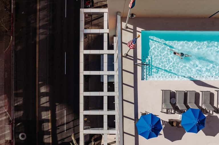 Aerial view of hotel pool with turquoise water, blue umbrellas, lounge chairs, and adjacent building