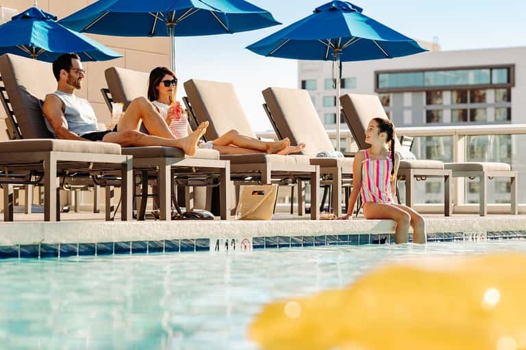 Family relaxing by resort pool with blue umbrellas and lounge chairs under clear sky