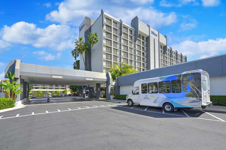 Modern hotel exterior with multi-story residential tower, covered entrance, and shuttle bus under clear blue sky