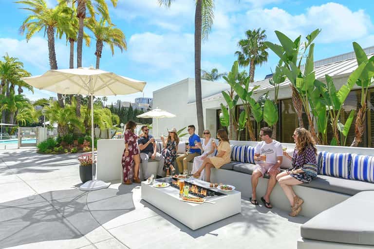 Modern poolside lounge area with white umbrellas, palm trees, and guests socializing on contemporary seating