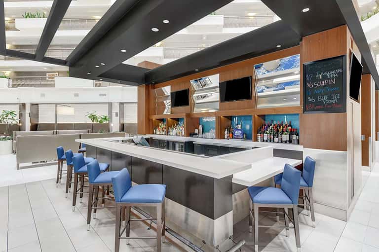 Modern hotel bar with blue bar stools, white countertop, wooden back panel with shelving, and black ceiling beams.