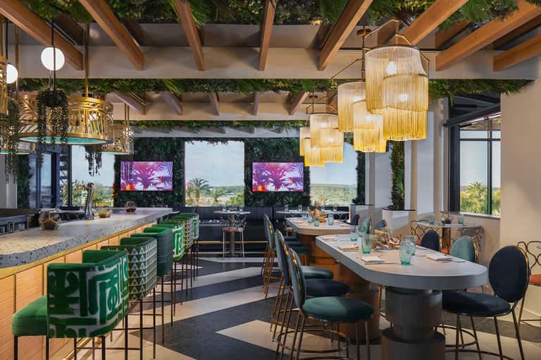 Modern restaurant with green velvet bar seating, wooden beams, hanging pendant lights, and panoramic windows overlooking landscaped grounds