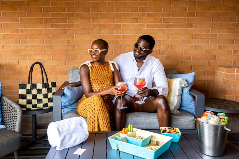 Couple sitting on a brick patio enjoying cocktails with snacks and a beach bag nearby