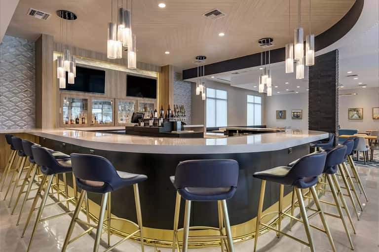 Contemporary hotel bar with curved black countertop, blue bar stools, pendant lighting, and wood finishes