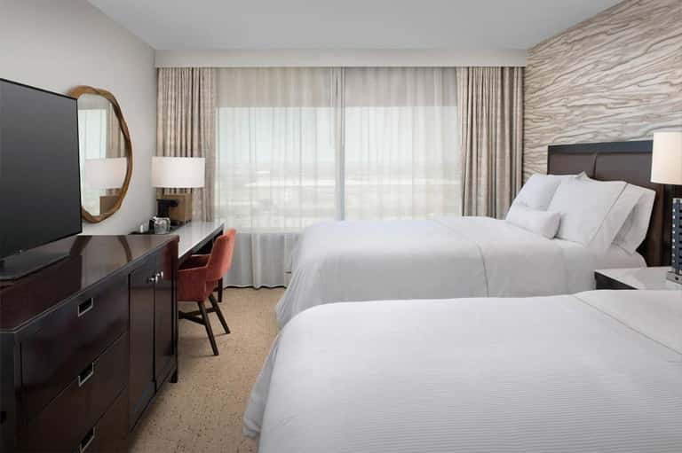 Modern hotel room with two queen beds, dark wood furniture, and floor-to-ceiling curtains