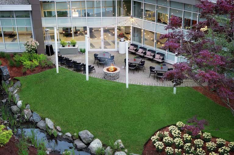 Modern outdoor patio with firepit, seating areas, manicured lawn, flowering plants, and large glass building.