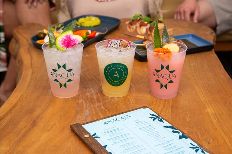 Three colorful cocktails with tropical garnishes on wooden table with appetizers