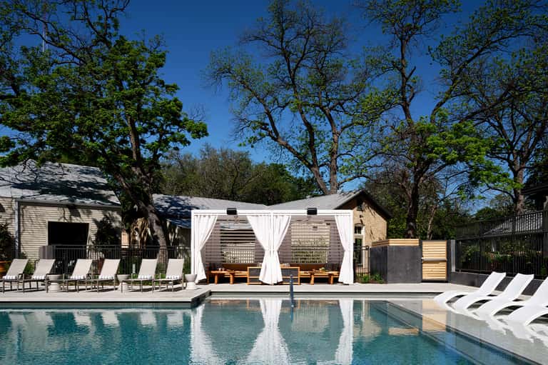 Modern rectangular pool with white lounge chairs and covered cabanas under mature oak trees