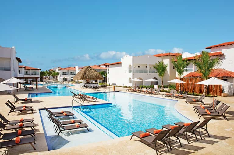 Resort pool area with loungers, white Mediterranean-style buildings, orange tile roofs, and palm trees under blue sky