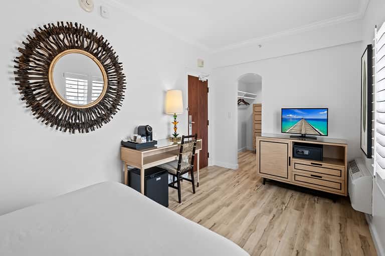 Hotel suite with queen bed, desk, TV, decorative sunburst mirror, and wood flooring