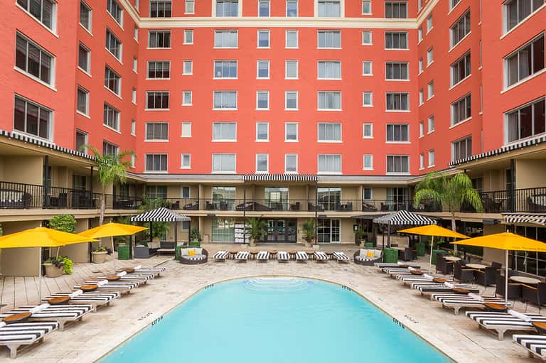 Coral-red courtyard building with striped lounge chairs, blue pool, yellow umbrellas, and black-and-white accent details