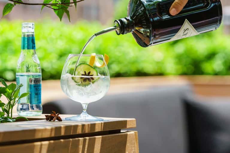Gin and tonic cocktail being poured into a glass with cucumber and star anise garnish on wooden table