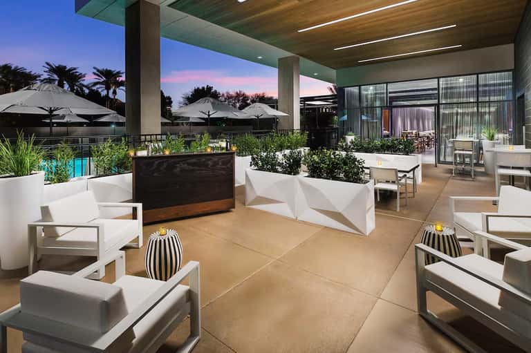 Modern rooftop terrace with white furniture, potted plants, umbrellas, and city skyline at dusk