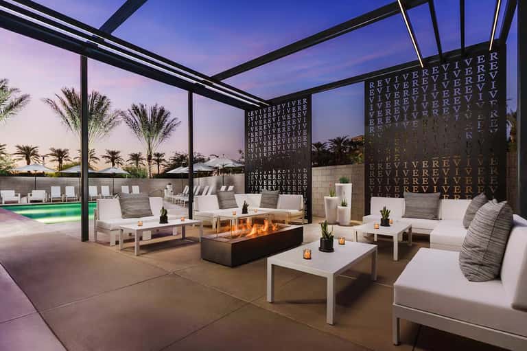 Modern rooftop lounge at dusk with fire pit, white seating, and palm trees under pergola structure
