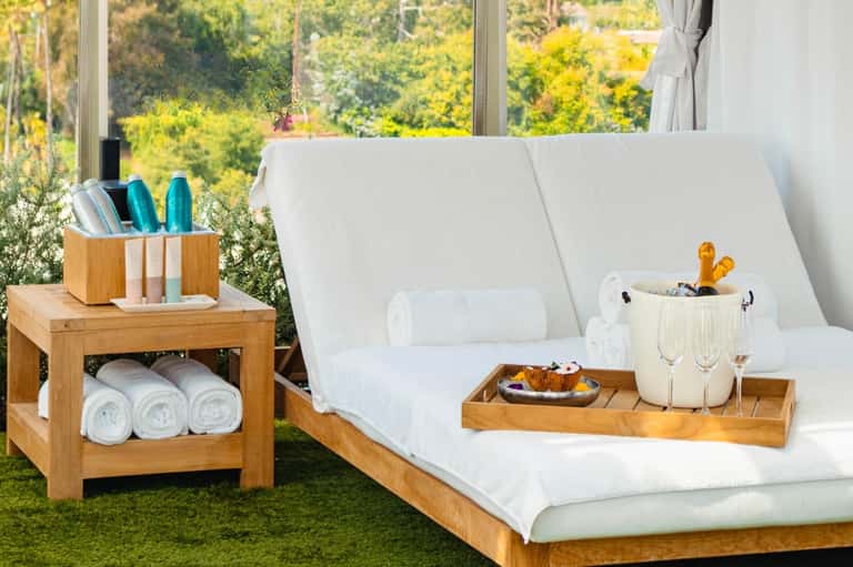 Spa lounge chairs with white cushions beside wooden side table holding rolled towels and amenities