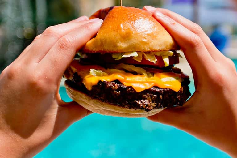 Hands holding a gourmet cheeseburger with golden bun and melted cheese against turquoise background