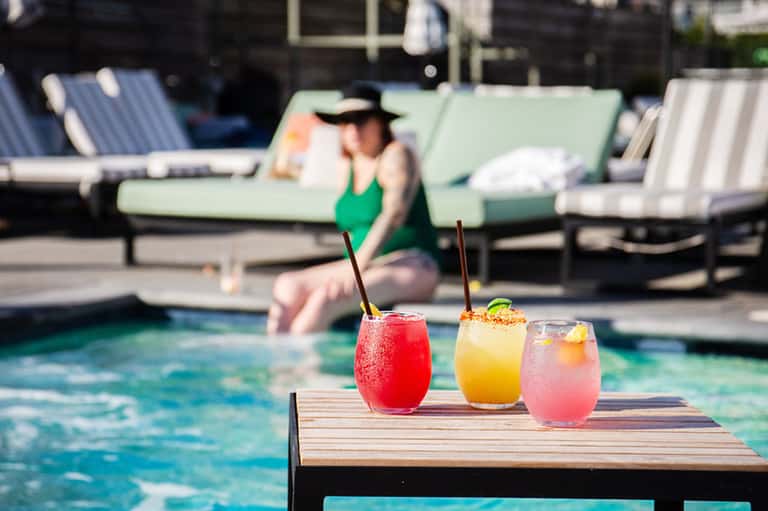 Colorful cocktails on poolside table with woman in green swimsuit relaxing in background