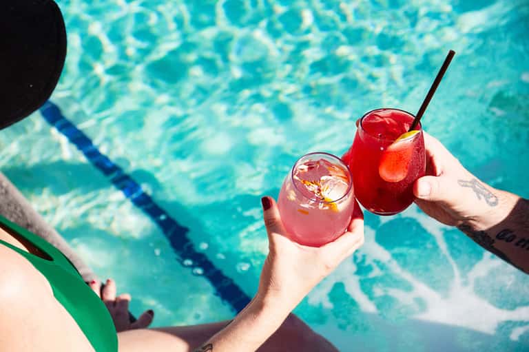 Two people holding colorful cocktails by a swimming pool with lane dividers