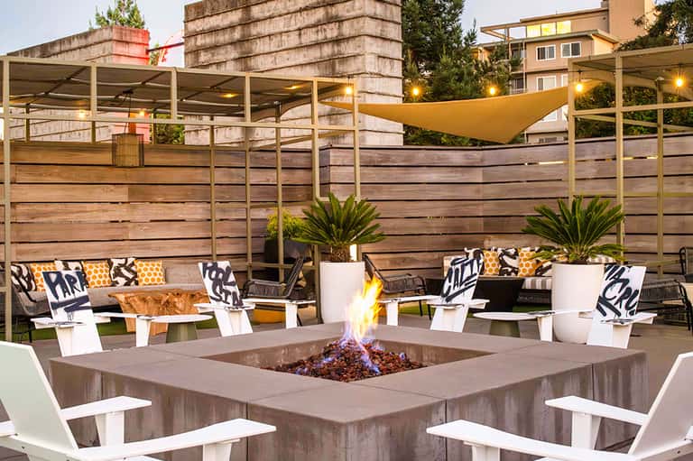 Evening rooftop lounge with fire pit, wooden privacy screens, yellow sail shade, and modern seating areas