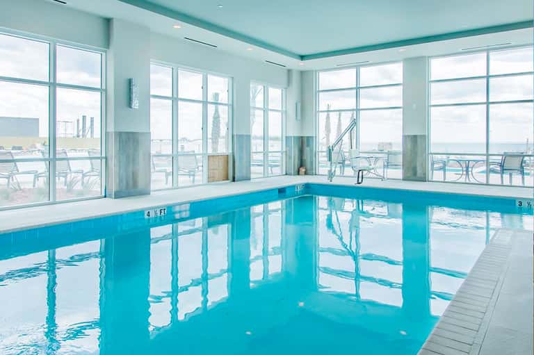 Bright indoor swimming pool with floor-to-ceiling windows overlooking an outdoor deck with lounge chairs