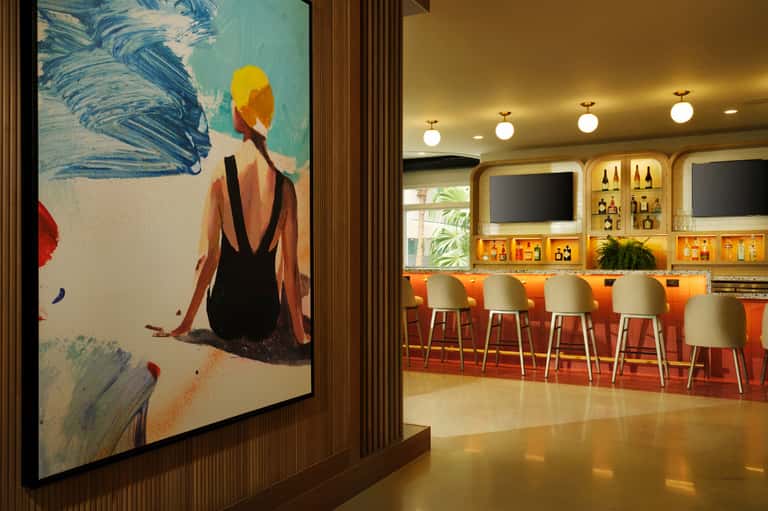 Modern hotel bar lounge with orange seating, illuminated shelving, and contemporary artwork featuring a beach scene