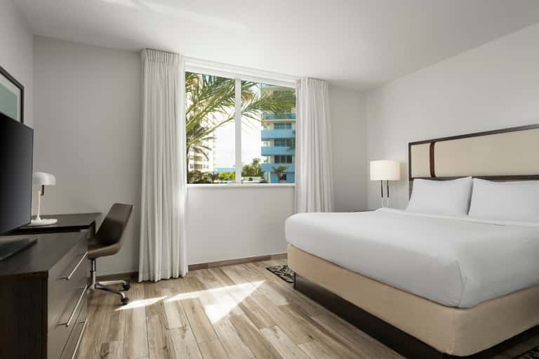 Modern hotel bedroom with ocean view through large windows, white bedding, dark wood desk, and light wood flooring