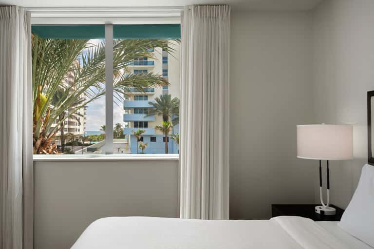 Beachfront hotel room with ocean view through large windows, palm trees visible, white bedding, and minimalist décor