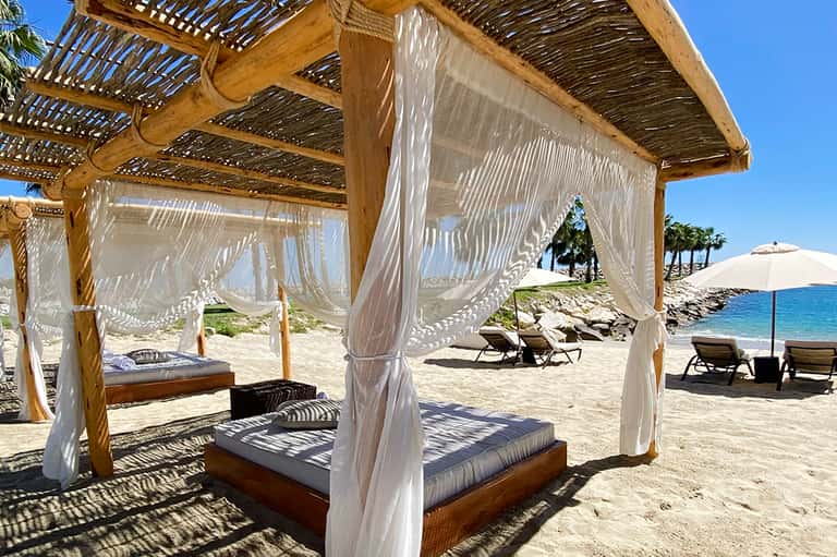 Beachfront cabanas with white curtains and wooden pergolas overlooking the ocean and sand