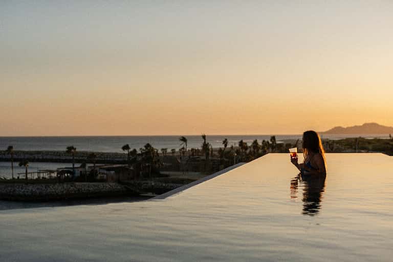 Woman in infinity pool at sunset overlooking coastal landscape with palm trees