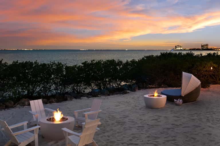 Beach bonfire setup at sunset with white Adirondack chairs and ocean view